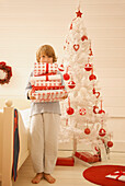 Junge steht an einem Weihnachtsbaum und hält einen Stapel Geschenke in der Hand