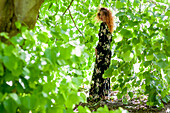 Frau in geblümtem Kleid zwischen Blättern im Wald stehend