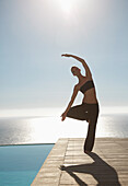 Junge Frau übt Yoga an einem Swimmingpool mit dem Meer im Hintergrund