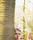 Mädchen versteckt sich hinter einem Baum, trägt indianischen Federschmuck und hält Pfeil und Bogen in der Hand, Nahaufnahme