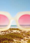 Ausschnittansicht einer rosafarbenen Sonnenbrille mit Strand im Hintergrund