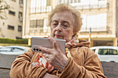 Ältere Frau sitzt auf einer Stadtbank und benutzt ihr Smartphone