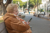 Ältere Frau sitzt auf einer Bank auf dem Bürgersteig und benutzt ihr Smartphone