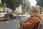 Ältere Frau sitzt auf einer Parkbank in der Stadt und benutzt ihr Smartphone