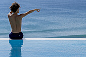 Junger Mann am Beckenrand sitzend und auf den Horizont zeigend am Meer