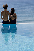 Rückansicht eines jungen Paares in Badekleidung, das am Rande eines Swimmingpools sitzt und auf das Meer blickt