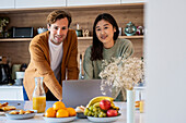 Adult couple looking at the camera while using laptop at kitchen counter