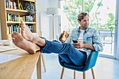 Junger erwachsener Mann ruht seine Füße aus, während er zu Hause mit seinem Smartphone arbeitet