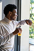 Seitenansicht eines erwachsenen Mannes, der am Fenster lehnt und Orangensaft trinkt