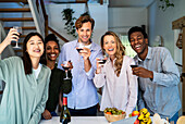 Group of friends looking at the camera toasting with wineglasses standing in kitchen