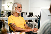 Erwachsener Mann mit langen grauen Haaren, der einen Computer benutzt, während er am Schreibtisch sitzt