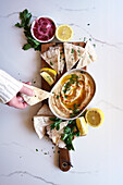 Chickpea hummus served with olive oil, pickled red onion, lemons, and flatbread on a white marble table background. Female hand dipping flatbread.