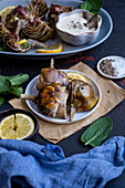 Roasted artichokes served on a white plate, accompanied by a bowl of dipping sauce, mint leaves and lemon slices photographed from front view.