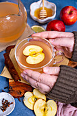 Hands holding a cup with apple tea, cloves, apples and honey in a small bowl around it
