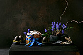 Hausgemachter traditioneller Oster-Panettone-Kuchen mit blauem Band, gefärbte schwarze Eier in Moos, blühende Muscari-Blumen auf schwarzem Holztisch. Traditionelle italienische Ostern backen, kopieren Raum