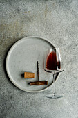 High angle view of a glass of dry red wine lying on a plate with corkscrew and cork on a concrete background