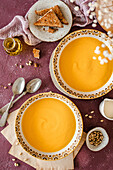 Two bowls of pumpkin soup, served with brown toast and garnished with cream, pine nuts and herbs on a purple background