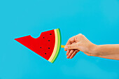 Unrecognisable person showing off a glowing paper watermelon with a bitten-off ice lolly on a stick against a blue background in the studio