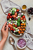 Flatlay eines griechischen Salats mit frischem Basilikum und Löffeln auf einem hellen Hintergrund mit einer Hand