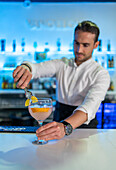 Focused male bartender using tongs to garnish a slice of orange on the rim of a cocktail glass while looking down and standing at the bar in front of blurred blue lights and bottles