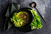 Top view of healthy celery cream soup in bowl with celery sticks served on black plate with black spoon and napkin against blurred dark background