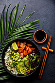 Lachs Poke Bowl Roher Fischsalat Asiatisches Trendgericht mit Sojabohnen, Edamame, Reis, Avocado und Salat in Schale auf tropischem Blatt und dunklem Hintergrund