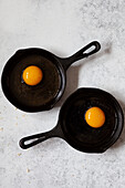 Zwei kleine gusseiserne Bratpfannen mit je einem rohen Ei darin