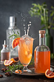 Action-Splash-Shot mit Eis, das in einen Blutorangen-Cocktail im Vintage-Glas fällt, mit Sirupflasche und Zitrus- und Rosmaringarnitur