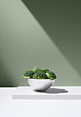 Fresh green broccoli growing in white bowl on plain table against gray background under bright ray of light