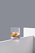 Transparent glass of whiskey on rocks with ice cubes placed on edge of white wooden table isolated on gray background