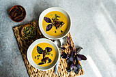 Top view of bowls of cream of pumpkin soup with basil herb, rye bread and seeds with spoons, bowl of seeds and leaves on brown tray