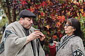 Serious mature couple in traditional Mapuche clothing with thermos flask and mug drinking hot yerba mate in the autumn garden in Temuco
