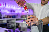 Unrecognisable male bartender pouring alcohol from a bottle into a jigger while standing and working at the bar counter in front of a blurred shop