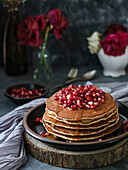 Stapel von Pfannkuchen mit Granatapfel, Karamell und roten Blumen auf einem schwarzen Teller und einem dunklen Hintergrund