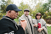Happy mature men and women in traditional dress chat and drink mate while spending time together on a lawn in the park