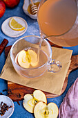 Apple tea is poured into a glass cup, apple slices, cloves, lemon slices, honey around it