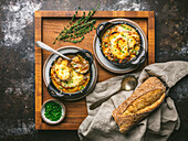 Two bowls of onion soup in cast iron bowls with melted cheese, spoon in bowl, on wooden tray with bread and chives