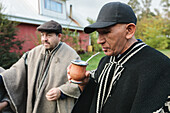 Nachdenklicher älterer Mann in traditioneller Mapuche-Kleidung und Baskenmütze, der wegschaut, während er steht und heißen Tee vor einem unscharfen Hintergrund in Temuco trinkt
