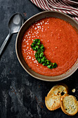 Gazpacho garniert mit Petersilie in einem Teller mit gewelltem Rand. Auf dem Tisch befinden sich rote Paprika, Tomaten, Knoblauch, Croutons und ein Löffel. Ansicht von oben