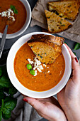 Eine Frau hält eine Schüssel Tomatensuppe mit Feta-Käse und Basilikumblättern und ein gegrilltes Käsesandwich in den Händen