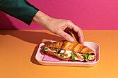 Cut-off, unrecognisable person eating a delicious croissant with ham, cheese and rocket on a plate in front of a colourful background