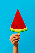Crop unrecognisable person demonstrating bright paper watermelon popsicle against blue background in studio
