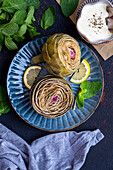 Steamed artichokes garnished with lemon slices and fresh mint leaves on a dark blue plate