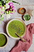 Flatlay einer Schüssel mit grüner Gemüsesuppe auf einer gefliesten Fläche mit Blumen