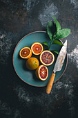 Blood oranges, halved and whole with citrus leaves, in a blue ceramic bowl with knife on a dark background