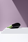 Vivid image of black eggplant with green cap lying on white stepped slab against purple background under bright light