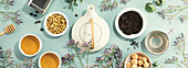 A white porcelain teapot, two cups, suga and assortment of dry tea in white ceramic bowls on blue background. Top view. Banner