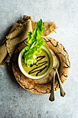 Healthy cream of celery soup in a bowl with celery stalks, served on a wooden plate with old spoons on a grey table next to a napkin