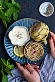 Woman serving steamed artichokes with a bowl of dipping sauce on a dark blue plate