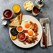 Ein komplettes englisches Frühstück mit Spiegeleiern, Würstchen, Speck, Blutwurst, Bohnen, Toasts und Tee auf grauem Betonhintergrund